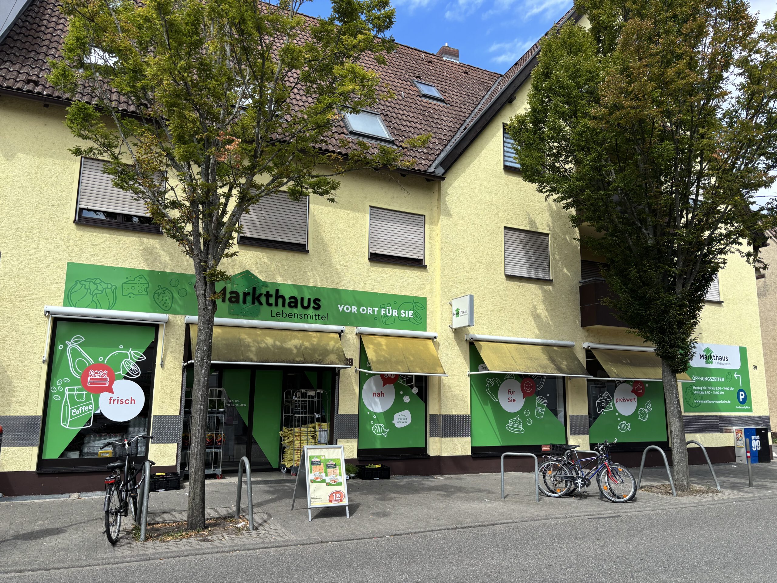 KI-basierte Bildbeschreibung. Ein kleines Lebensmittelgeschäft namens Markthaus mit grüner Beschilderung und Markisen befindet sich im Erdgeschoss eines gelben Gebäudes. Draußen sind Fahrräder abgestellt, und zwei Bäume säumen den Gehweg davor.