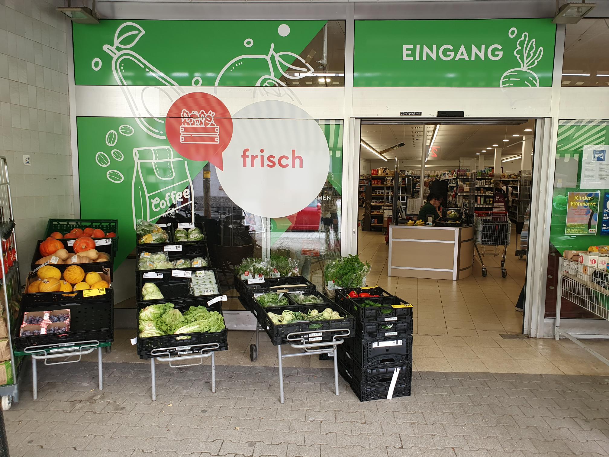 KI-basierte Bildbeschreibung. Frisches Gemüse und Kräuter in schwarzen Kisten vor dem Eingang eines deutschen Supermarkts. Über den Glastüren steht das Wort Eingang, und im Inneren des Ladens ist eine Kassiererin zu sehen.