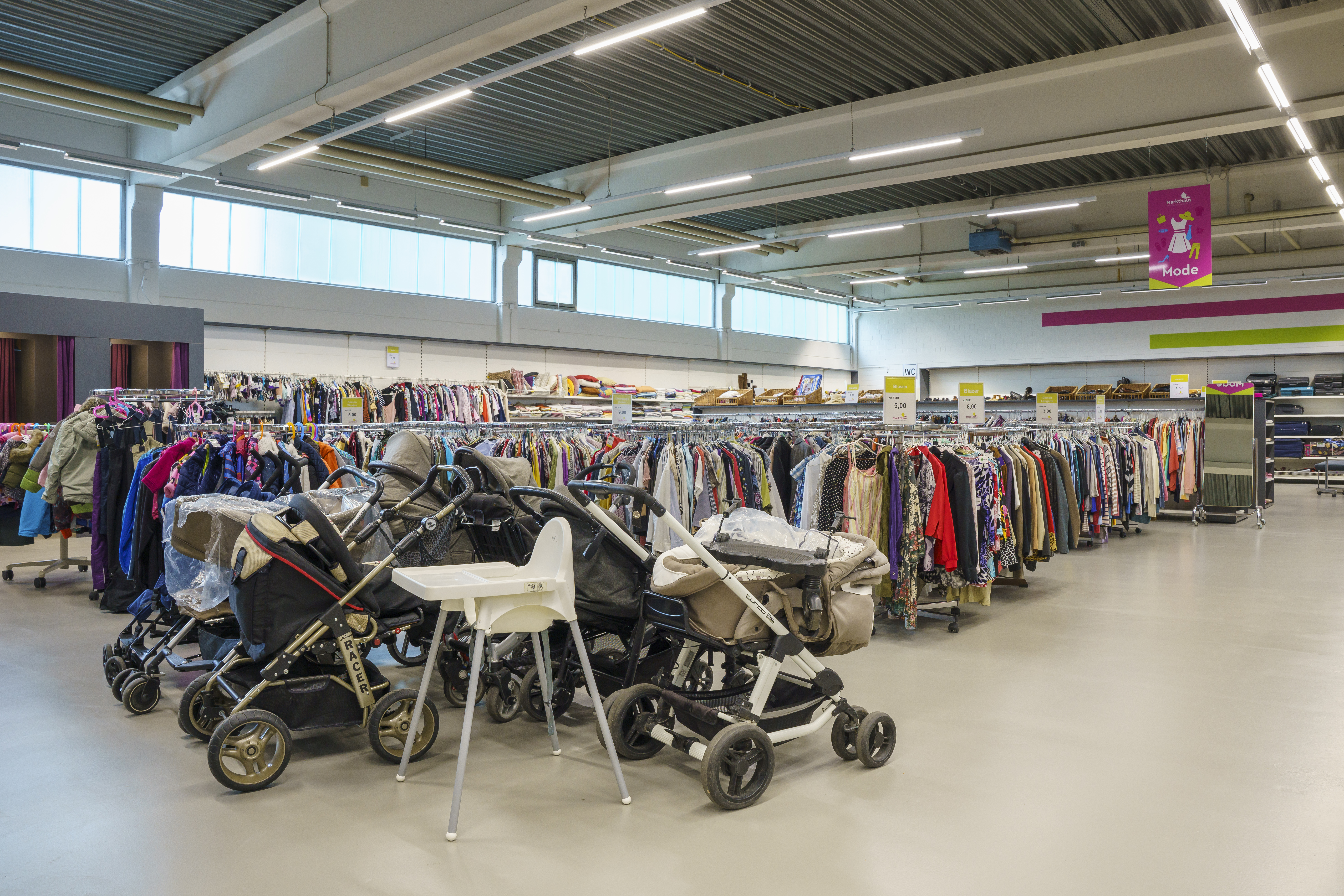 KI-basierte Bildbeschreibung. Reihen von Kleiderständern in einem hell erleuchteten Secondhand-Laden, mit mehreren Kinderwagen und einem weißen Hochstuhl im vorderen Teil des Ladens.