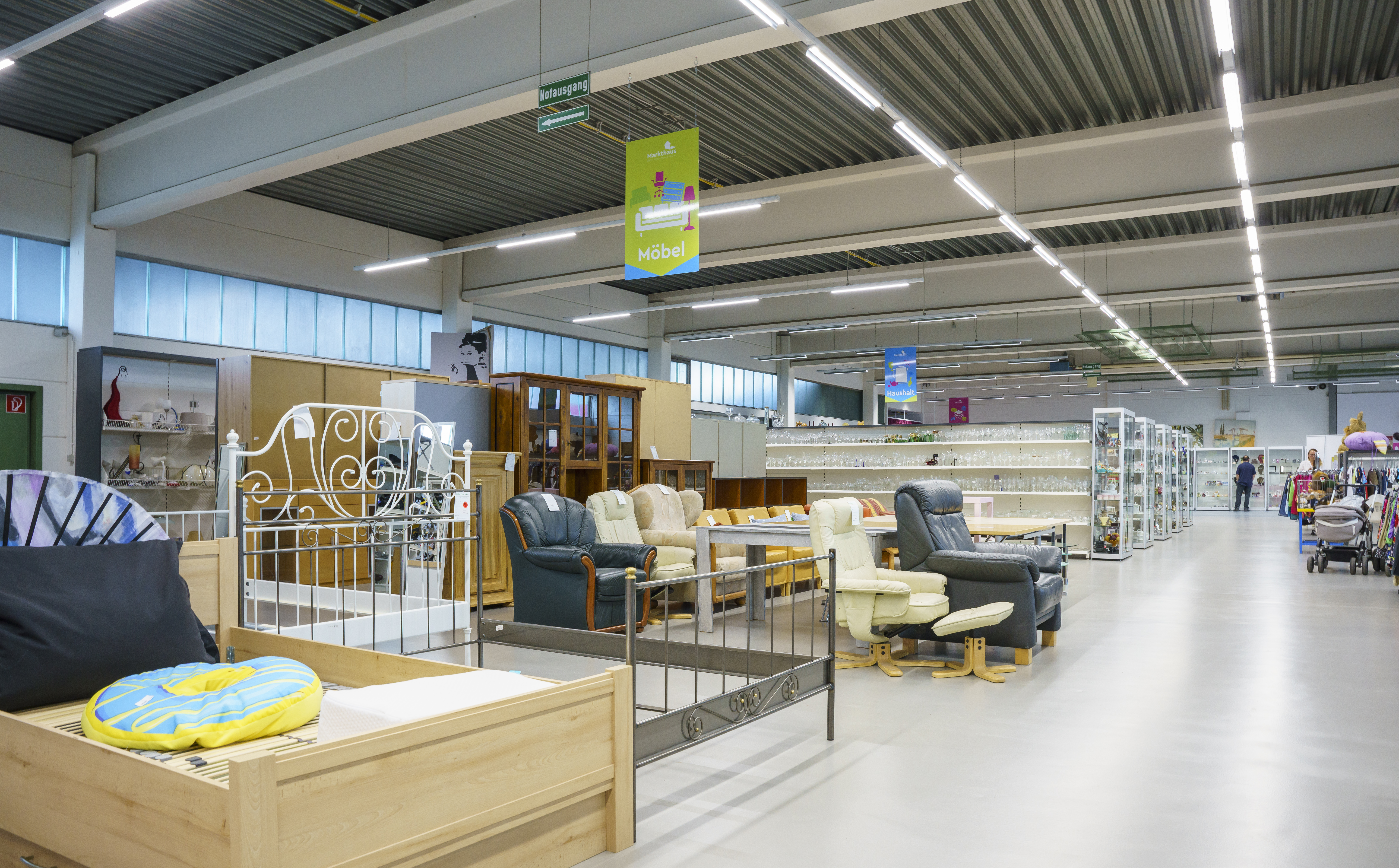 KI-basierte Bildbeschreibung. Ein geräumiger Secondhand-Laden mit ordentlich angeordneten Secondhand-Möbeln, darunter Stühle, Tische, Betten und Regale. Von der Decke hängen Leuchtstoffröhren, und Schilder in deutscher Sprache kennzeichnen die verschiedenen Abteilungen.