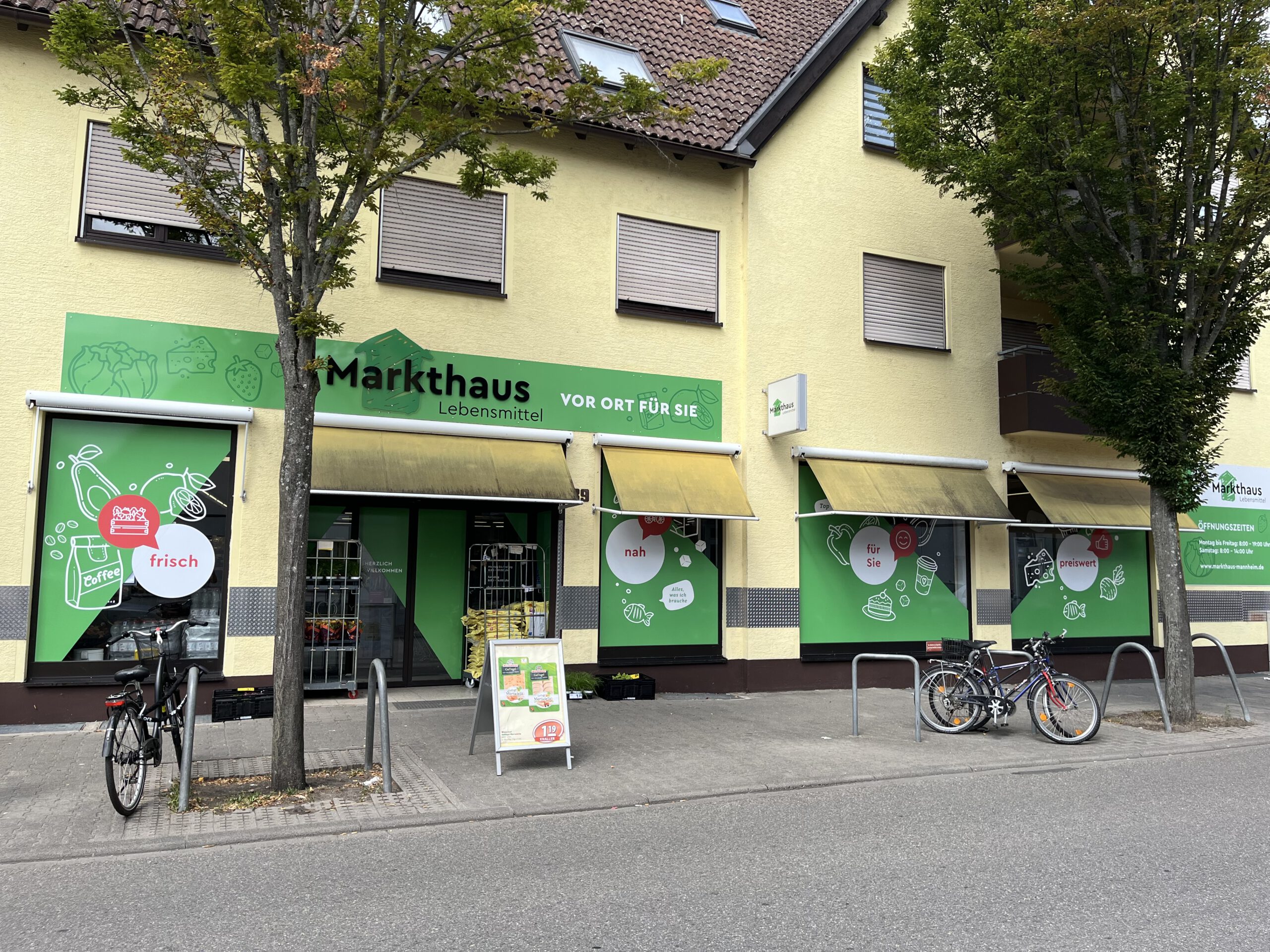 KI-basierte Bildbeschreibung. Ein kleines Lebensmittelgeschäft namens Markthaus Lebensmittel mit grünen Schildern liegt in einer ruhigen Straße. Vor dem Geschäft sind Fahrräder abgestellt, und der Gehweg ist von Bäumen gesäumt. Gelbe Markisen bedecken die Fenster und den Eingang.