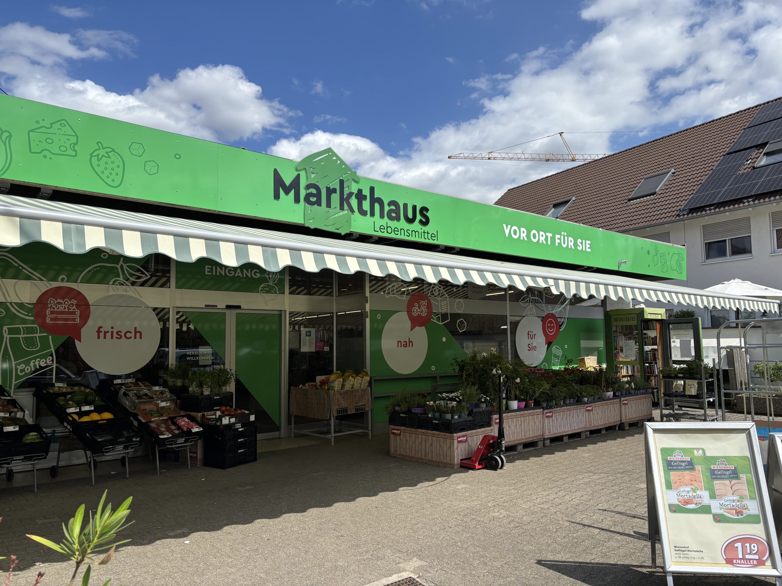 KI-basierte Bildbeschreibung. Eine grüne Markise verdeckt den Eingang des Markthauses Lebensmittel, in dem frische Produkte und Pflanzen angeboten werden. Schilder und Banner werben für verschiedene Artikel, und im Hintergrund sind Wohnhäuser zu sehen.