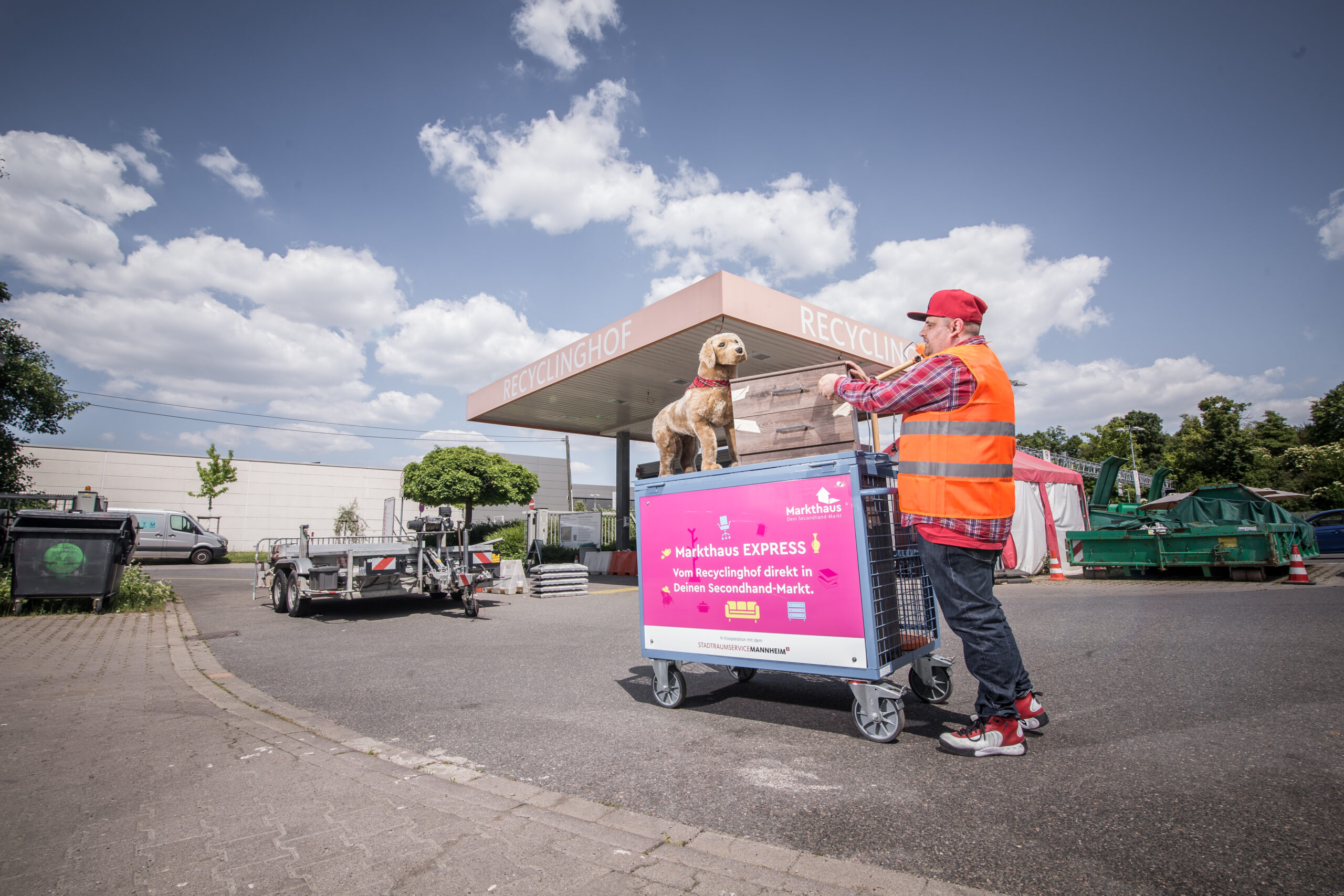 KI-basierte Bildbeschreibung. Eine Person in orangefarbener Sicherheitsweste und roter Mütze steht an einem Recyclingzentrum und schiebt einen Wagen mit Kisten und einem Hund, der darauf sitzt. Der Himmel ist blau und wolkenverhangen, und im Hintergrund sind Recyclingtonnen zu sehen.