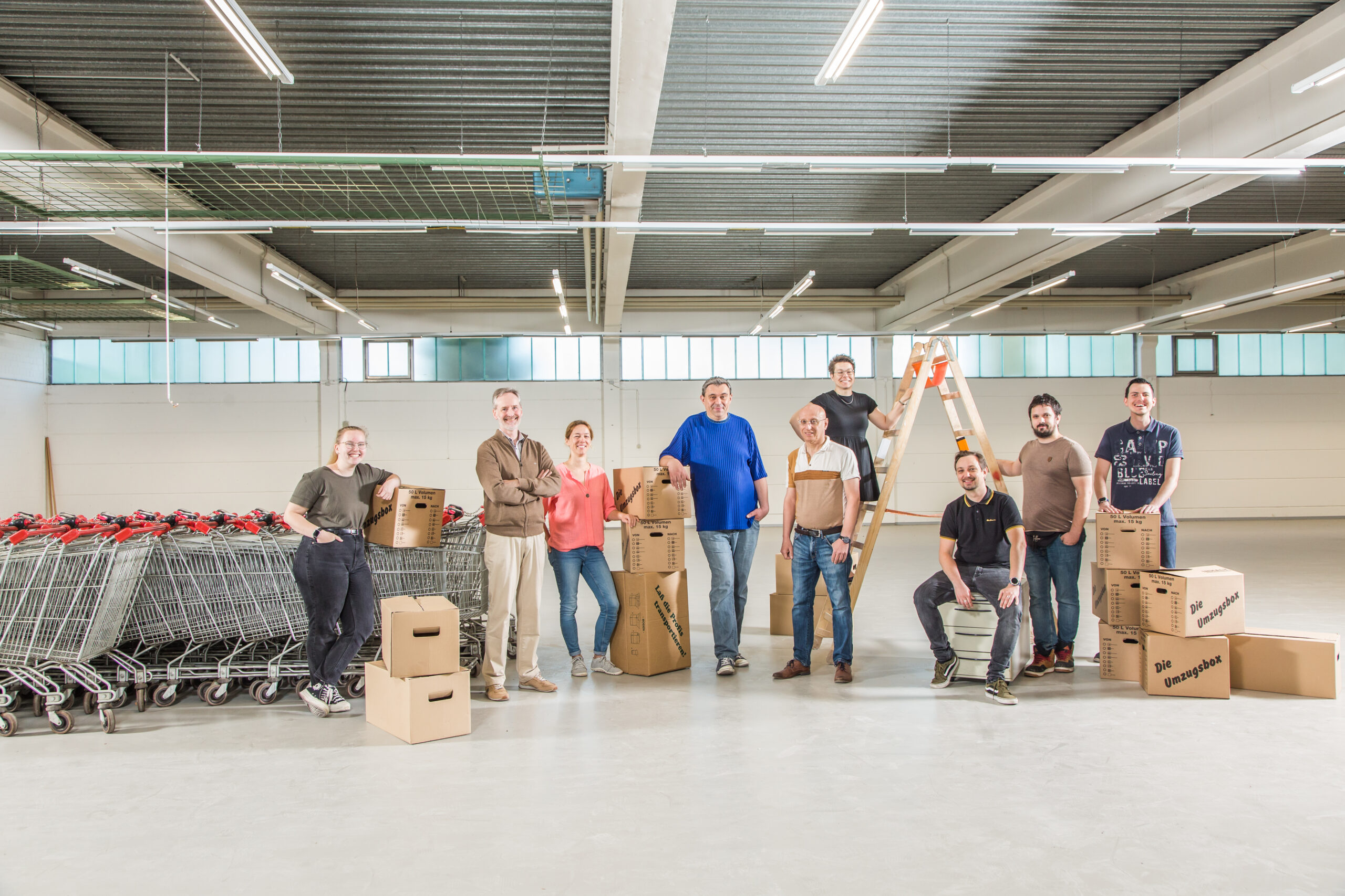 KI-basierte Bildbeschreibung. Eine Gruppe von Menschen steht und sitzt in einem großen, leeren Lagerhaus mit Einkaufswagen und Pappkartons, von denen einige gestapelt sind und einige gehalten werden, und einer Person auf einer Leiter im Hintergrund.