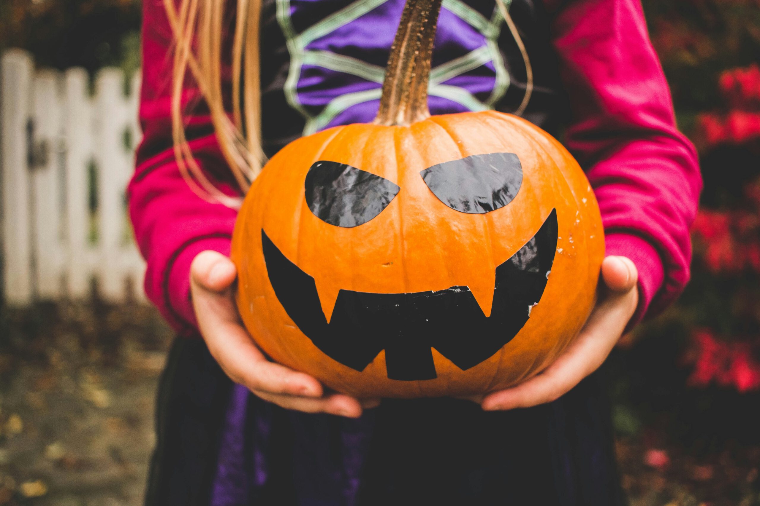KI-basierte Bildbeschreibung. Ein Kind in einem Kostüm hält einen geschnitzten Kürbis, der mit einem schwarzen, bedrohlichen Gesicht für Halloween verziert ist. Der Kürbis hat scharfe Zähne und schräge Augen; der Hintergrund ist unscharf mit einem weißen Zaun und Herbstfarben.
