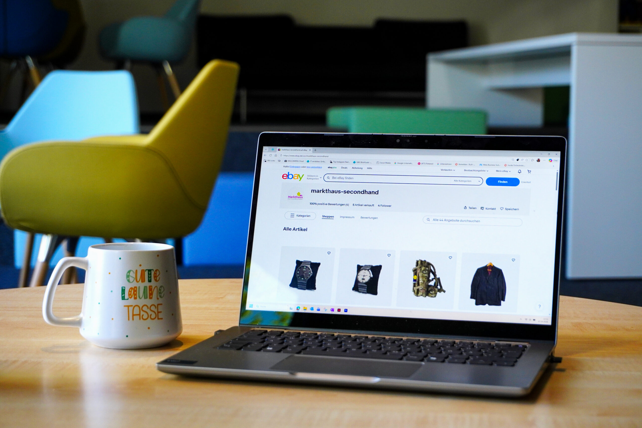 KI-basierte Bildbeschreibung. Ein Laptop auf einem Holztisch zeigt eine eBay-Seite für Secondhand-Kleidung an. Daneben steht eine weiße Tasse mit buntem Text. Im Hintergrund befinden sich moderne Stühle und ein Tisch in einem hellen Büroraum.