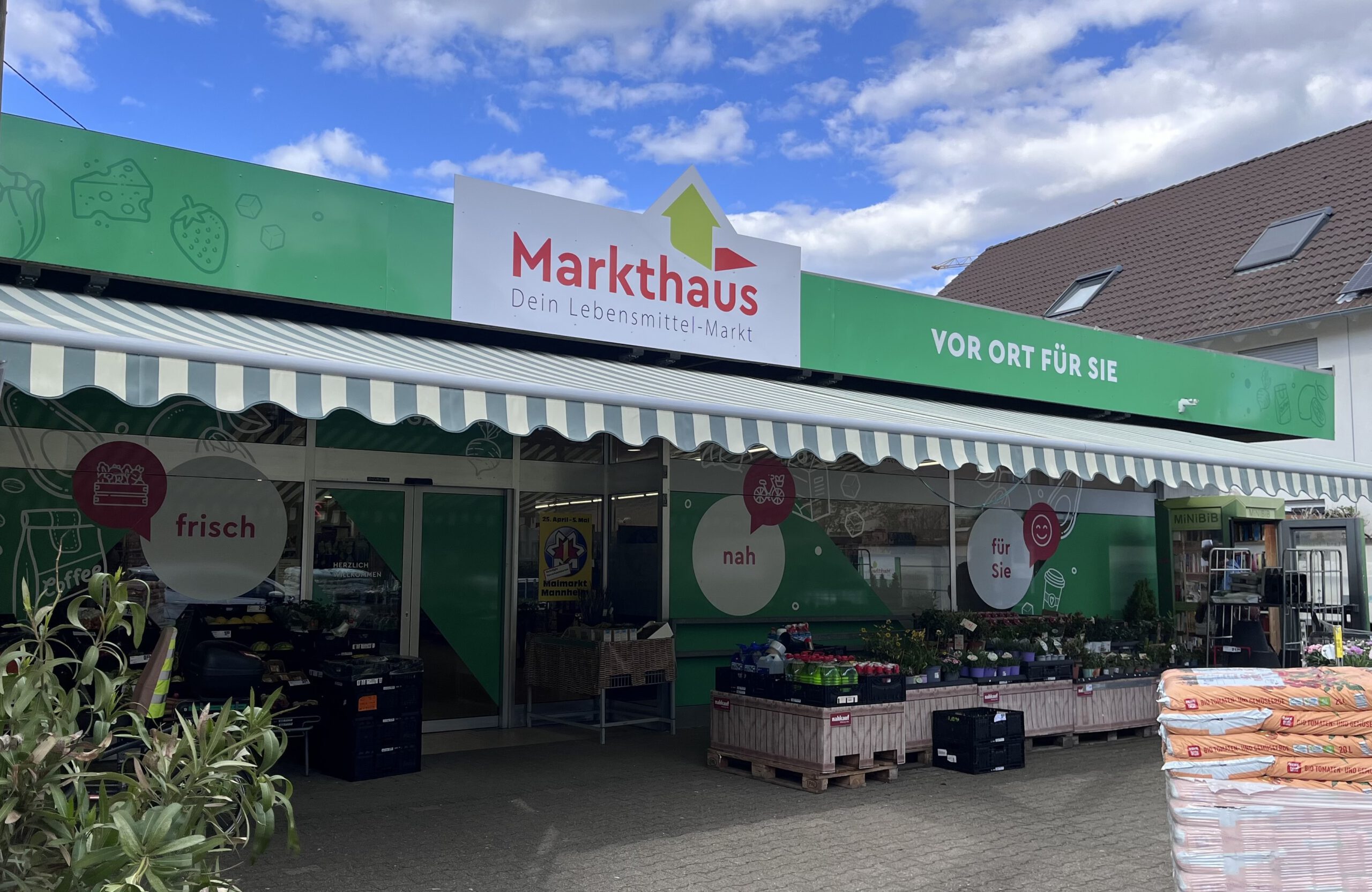 KI-basierte Bildbeschreibung. Ein grün-weißes Schaufenster von Markthaus, einem Lebensmittelgeschäft in Deutschland, mit frischen Produkten und Pflanzen unter einer Markise. Der Himmel ist teilweise bewölkt und ein benachbartes Haus ist zu sehen.
