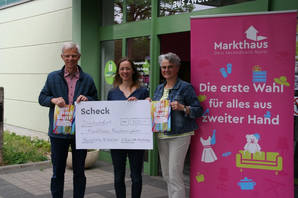 KI-basierte Bildbeschreibung. Drei Personen stehen draußen, lächelnd, mit einem großen Scheck über 300 Euro und bunten Geschenktüten in der Hand. Neben ihnen hängt ein leuchtend rosa Banner für das Markthaus, einen Secondhand-Markt.