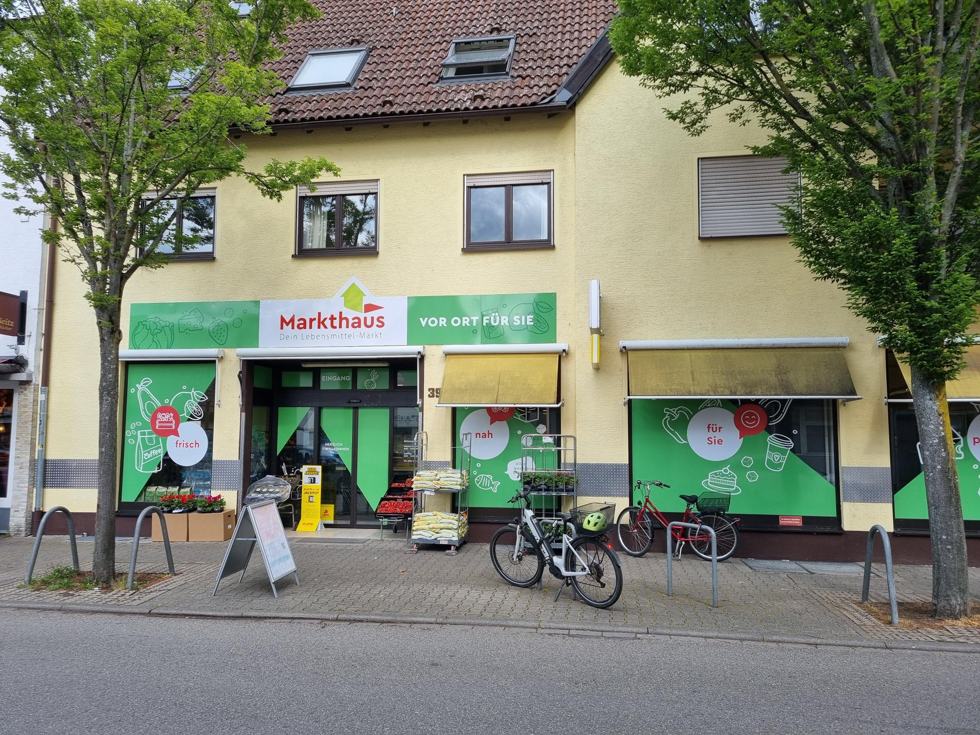 KI-basierte Bildbeschreibung. Ein kleines Lebensmittelgeschäft namens Markthaus mit grüner Beschilderung, Fahrrädern, die draußen geparkt sind, Tüten mit Waren am Eingang und Bäumen auf dem Bürgersteig vor einem gelben Gebäude.