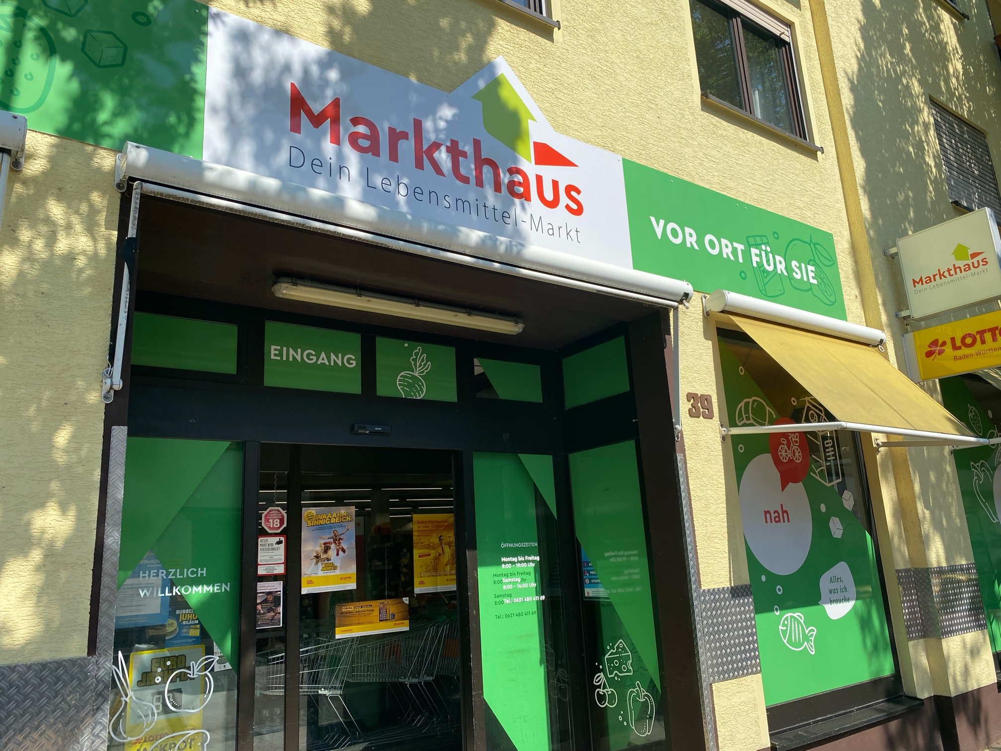 KI-basierte Bildbeschreibung. Der Eingang eines Lebensmittelgeschäfts namens Markthaus mit grüner Beschilderung, Glastüren und einer gelben Markise. Auf den Schildern steht "Dein Lebensmittel-Markt" und "Vor Ort Für Sie". Im Inneren sind Einkaufswagen zu sehen.