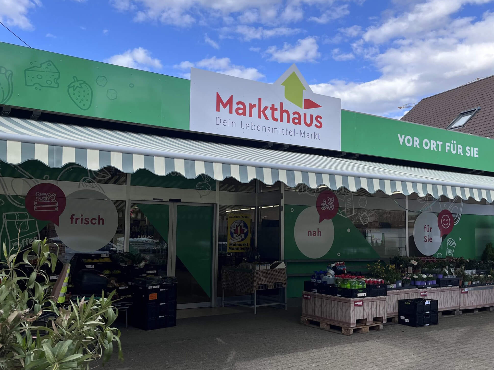 KI-basierte Bildbeschreibung. Außenansicht eines kleinen Lebensmittelladens namens Markthaus mit grüner Beschilderung, frischen Produkten in Kisten draußen und einer Markise über dem Eingang. Es gibt Schilder mit deutschen Wörtern und einen teilweise bewölkten Himmel darüber.