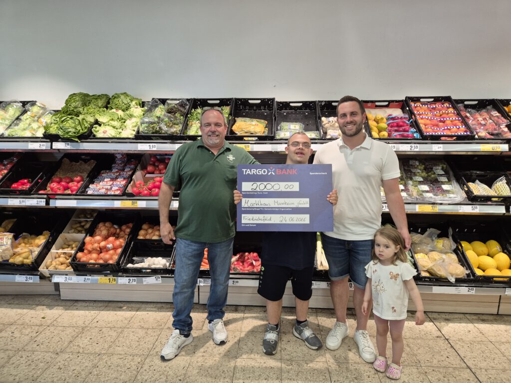 KI-basierte Bildbeschreibung. Drei Erwachsene und ein junges Mädchen stehen im Obst- und Gemüseregal eines Supermarkts. Die Erwachsenen lächeln und halten einen großen Symbolscheck der Targo Bank in den Händen. Hinter ihnen sind frisches Obst und Gemüse ausgestellt.